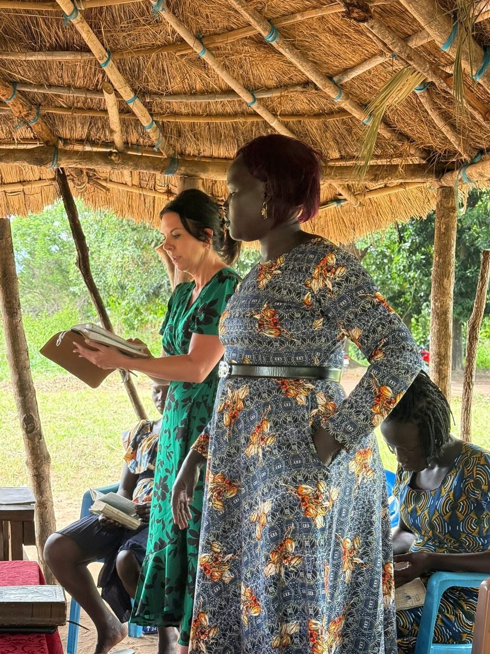 Jodi reading Scripture under a thatched shelter while a woman teaches beside her